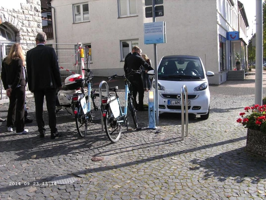 Polizisten kontrollieren ein geparktes, weißes Auto neben Fahrrädern auf einer gepflasterten Straße.