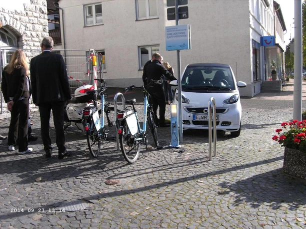 Polizisten kontrollieren ein geparktes, weißes Auto neben Fahrrädern auf einer gepflasterten Straße.