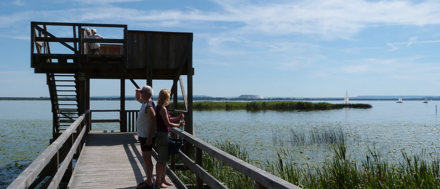 Aussichtsplattform Moorhütte Ein Paar genießt die Aussicht auf einem hölzernen Steg mit Plattform, umgeben von einem See und Segelbooten.