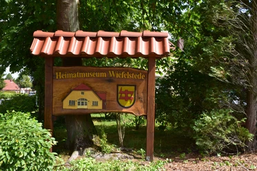 Heimatmuseum Holzschild mit dem Text "Heimatmuseum Wiefelstede" und einem Gebäude- und Wappenmotiv, umgeben von grüner Natur.