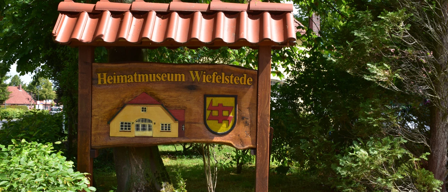 Heimatmuseum Holzschild mit dem Text "Heimatmuseum Wiefelstede" und einem Gebäude- und Wappenmotiv, umgeben von grüner Natur.