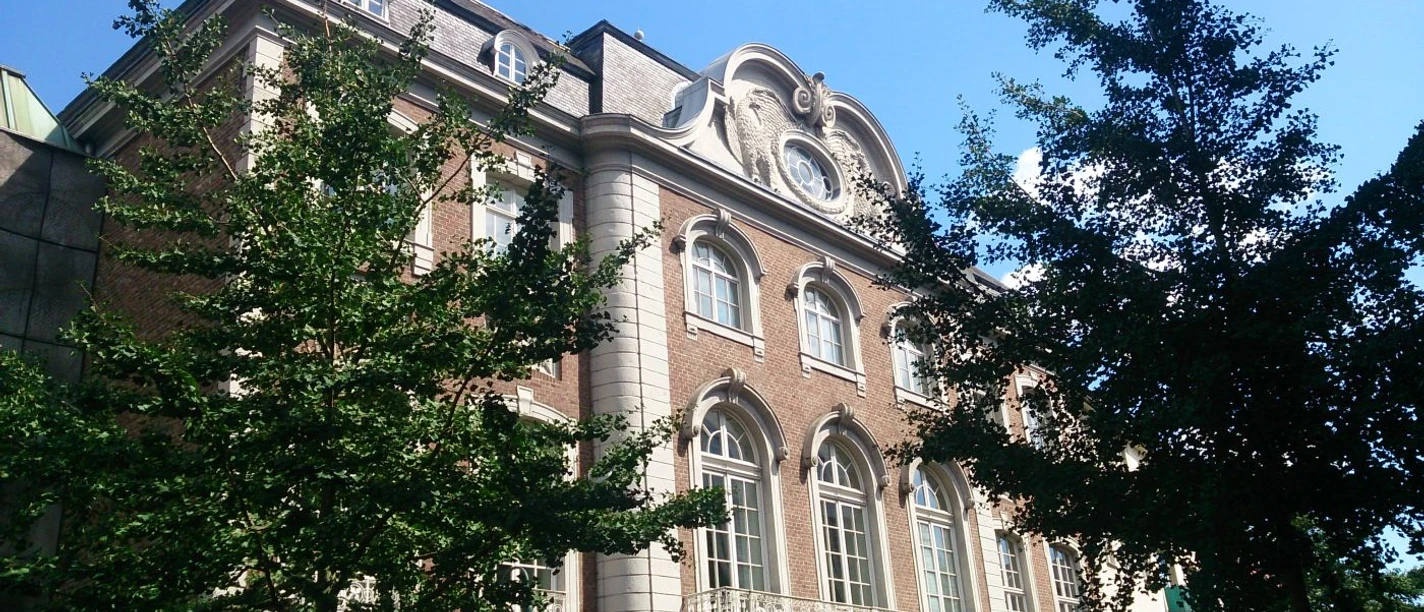 Altes Kurhaus Aachen Dreigeschossiges Gebäude im neoklassizistischen Stil, umgeben von grünen Bäumen und einem strahlend blauen Himmel.