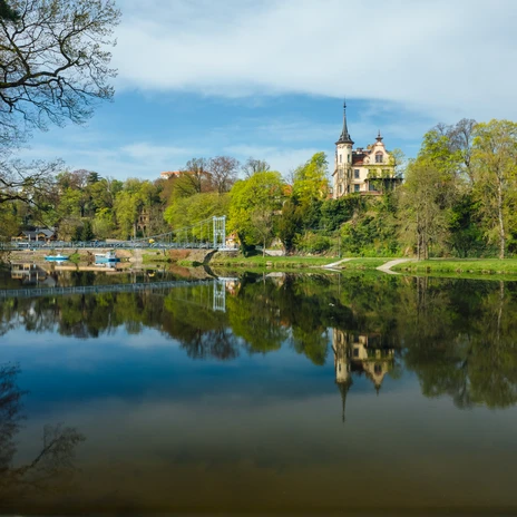 View of a historic bridge and a castle, surrounded by green nature and reflecting water.