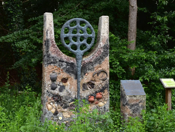 Hügelgrab Das Bild zeigt eine steinerne Skulptur mit kreisrunden und parallel angeordneten Elementen sowie einem angrenzenden Infotafel im Grünen.