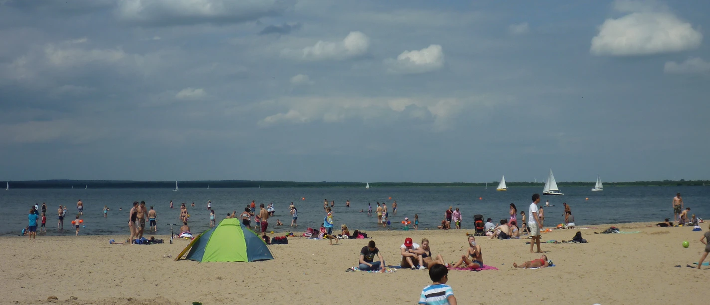 Een zonnig strand vol mensen met kleurrijke zwemkleding en parasols aan het Steinhuder Meer. Zeilboten glijden rustig over het water.