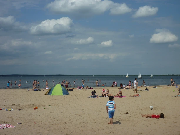 Badeinsel Steinhude Ein sonniger Strand voller Menschen mit bunten Badeanzügen und bunten Schirmen am Steinhuder Meer. Segelboote gleiten ruhig auf dem Wasser.