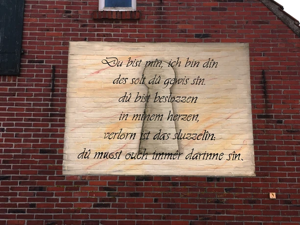 2. Mauergedicht - Meiners, Sögel Mittelalterliches Liebesgedicht auf gemalter Tafel an roter Backsteinwand mit schmalem Fenster oben.
