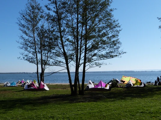 Am Ufer stehen bunte Kites, während Menschen das sonnige Wetter am Wasser genießen.