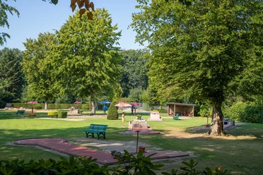 minigolf-uebersicht Ein weitläufiger Minigolfplatz, umgeben von Bäumen, mit bunten Sonnenschirmen und Bänken im Grünen.