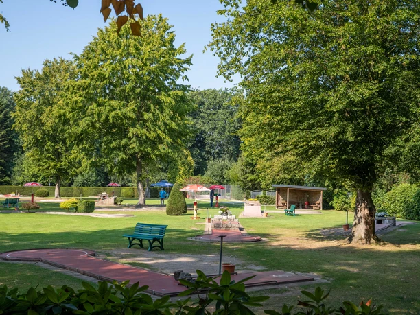 minigolf-uebersicht Ein weitläufiger Minigolfplatz, umgeben von Bäumen, mit bunten Sonnenschirmen und Bänken im Grünen.