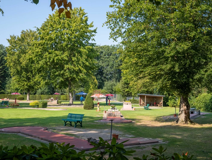 minigolf-uebersicht Ein weitläufiger Minigolfplatz, umgeben von Bäumen, mit bunten Sonnenschirmen und Bänken im Grünen.