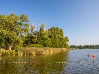 ufer-rostrup Ein sonniger See mit grünem Ufer und Bojen, umgeben von Bäumen unter klarem Himmel.