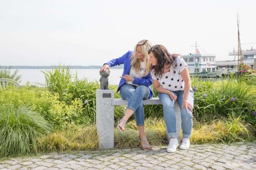 mainzelmaennchen Zwei Frauen sitzen auf einer Steinbank am See und berühren eine kleine Mainzelmännchen-Statue.