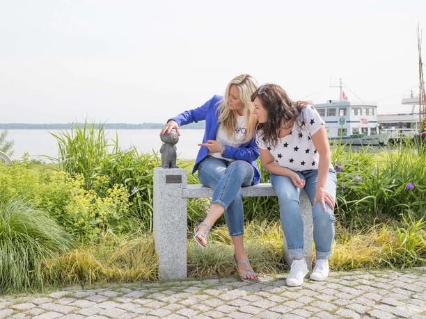 mainzelmaennchen Zwei Frauen sitzen auf einer Steinbank am See und berühren eine kleine Mainzelmännchen-Statue.