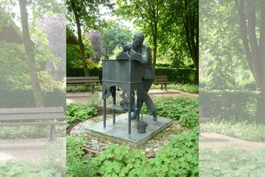 Albert Trautmann Denkmal Werlte Bronzestatue eines Mannes am Schreibpult, umgeben von Grünpflanzen und Parkbänken im Garten.