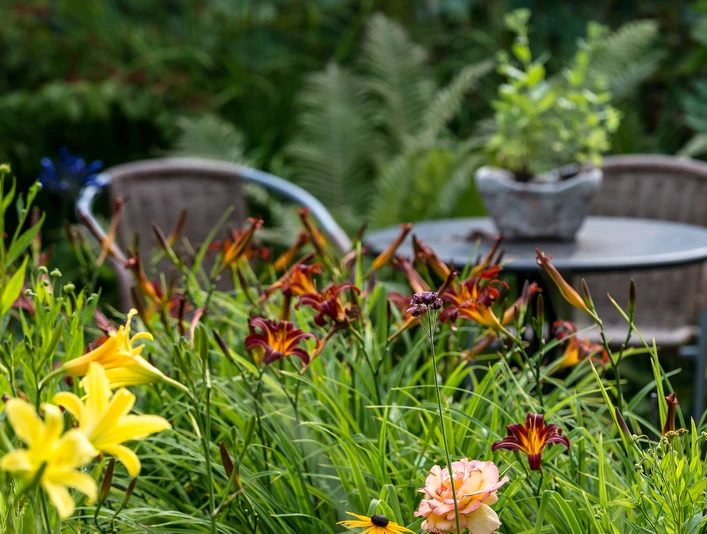 garten-stolle-wiefelstede-3_quer Ein idyllischer Garten mit bunten Lilien und einem runden Tisch vor zwei Rattanstühlen lädt ein.