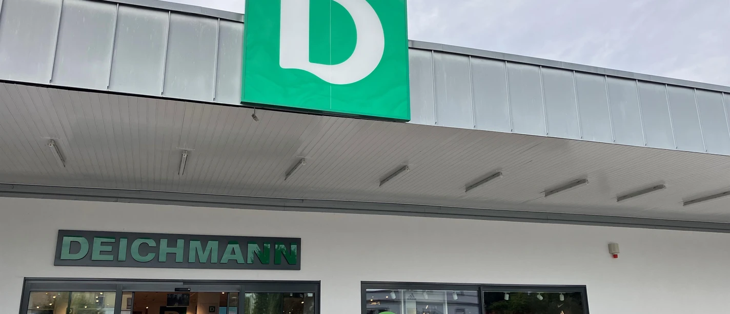 Eingang einer Deichmann-Filiale mit grünem Logo und parkenden Autos davor