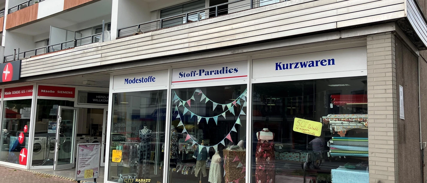 Schaufenster des Stoff-Paradies mit Stoffballen, Kleidern und Kurzwaren unter Wohnbalkonen