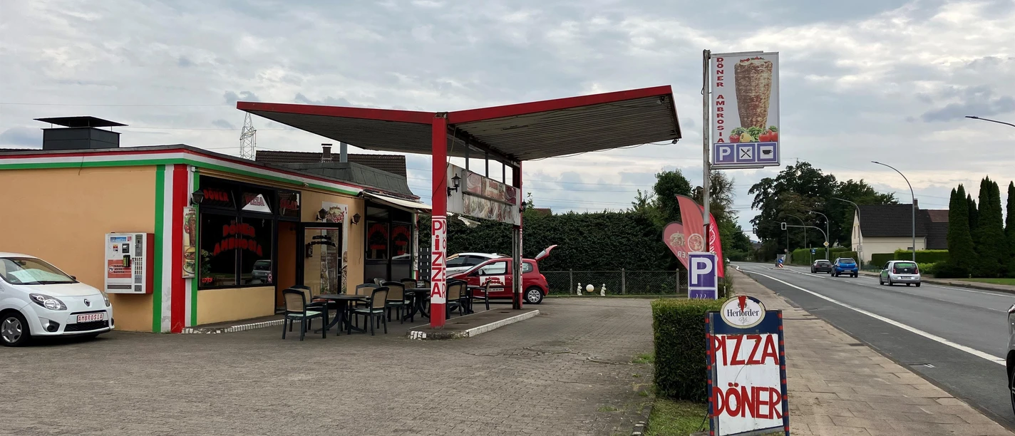 Döner Ambrosia mit überdachter Terrasse, Parkplatz und Straßenschild bei bewölktem Himmel