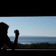 Aussichtsturm Ostufer Der dunkle Schatten einer Person, die eine Kamera hält, rahmt den weitläufigen und ruhigen Blick auf einen blauen See ein, während im Hintergrund zarte Hügel und Segelboote zu sehen sind.