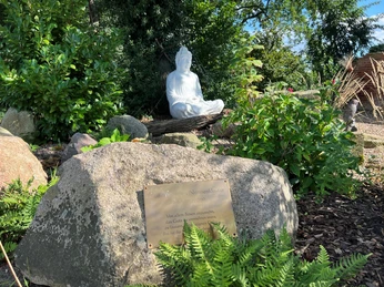 Buddha-Statue in einem grünen Garten, umgeben von Pflanzen und einem Stein mit Inschrift im Vordergrund.