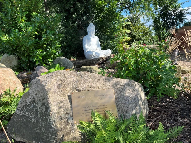 Buddha-Statue in einem grünen Garten, umgeben von Pflanzen und einem Stein mit Inschrift im Vordergrund.