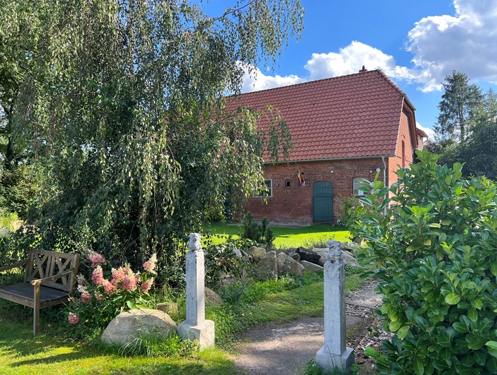 Rotes Backsteingebäude mit begrüntem Vorgarten, umgeben von Bäumen unter blauem Himmel.