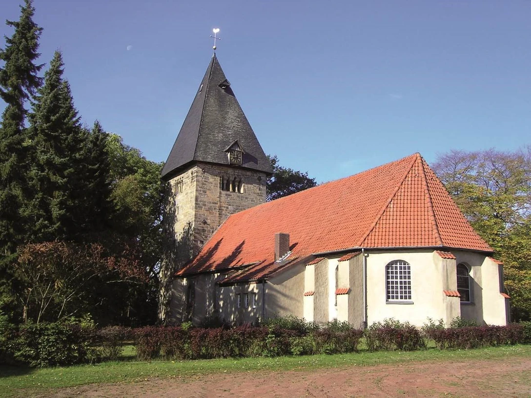 Kirche Basse Eine historische Kirche mit spitzem Turm, roten Ziegeldach und umgeben von Bäumen unter blauem Himmel.