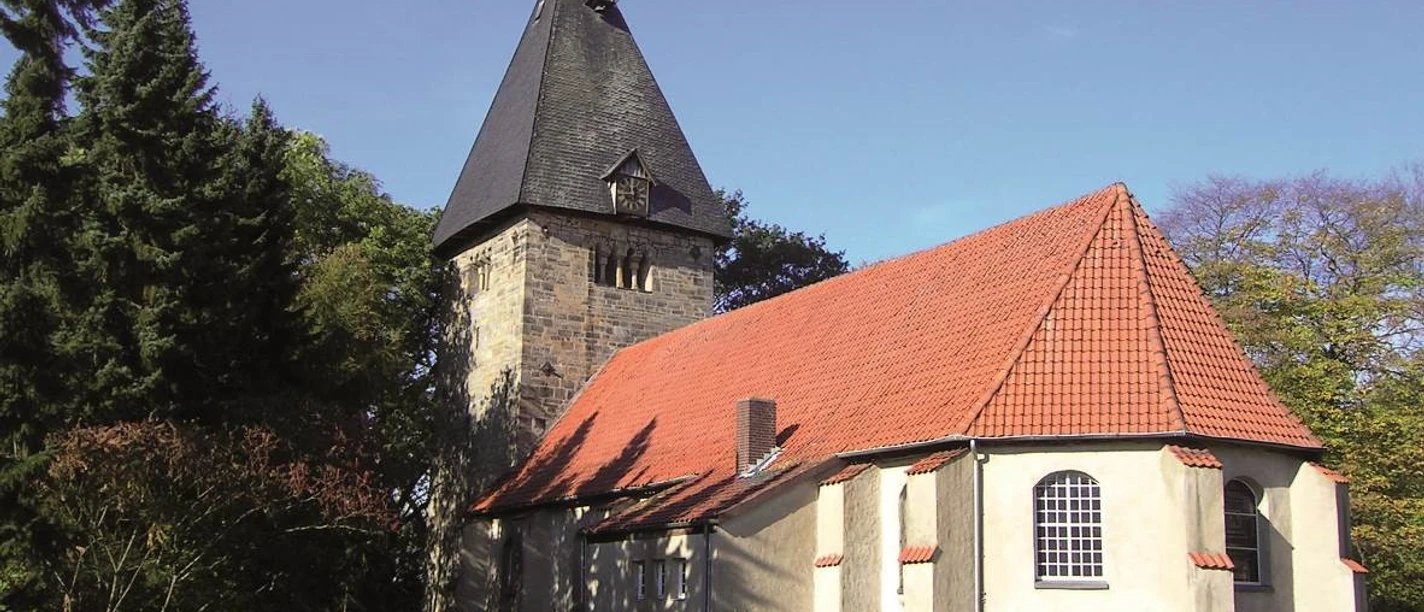 Kirche Basse Eine historische Kirche mit spitzem Turm, roten Ziegeldach und umgeben von Bäumen unter blauem Himmel.