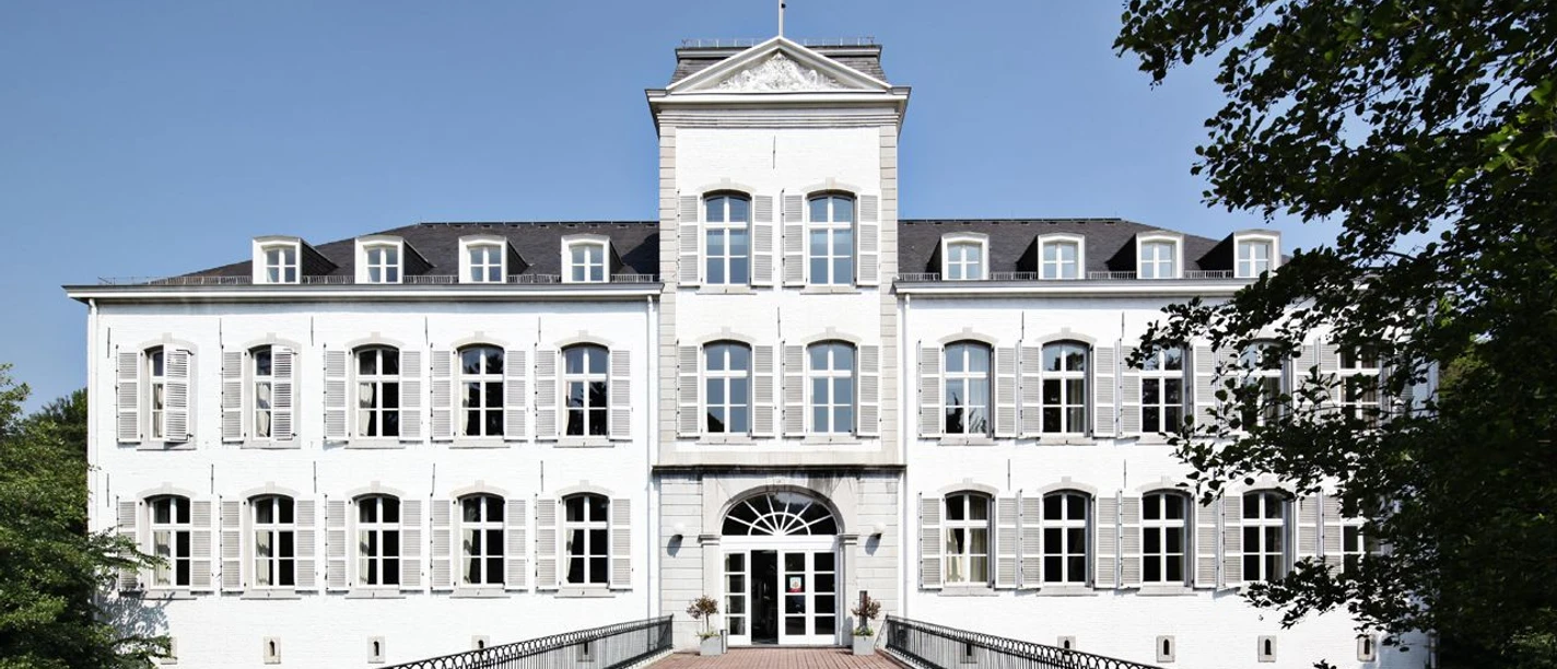 Schloss Rahe Aachen Weiße Fassade eines Schlosses unter klarem, blauem Himmel. Ein Weg führt über eine Brücke zum Vordereingang mit großen Fenstern und einem gepflegten Eingangsbereich.