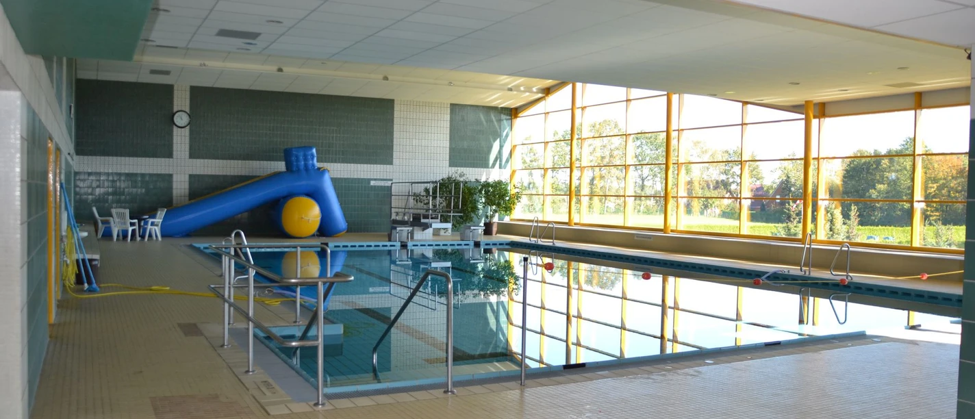 Hallenbad Werlte Innenansicht eines hellen Hallenbads mit großem Becken, blauer Rutsche und Blick ins Grüne.