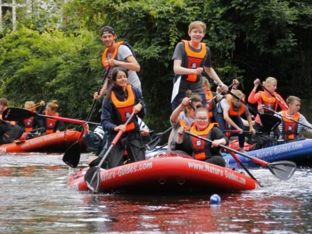 Jugendliche in orangen Rettungswesten paddeln auf großen roten SUP-Boards auf einem Fluss.