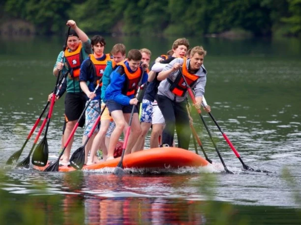 Acht junge Männer paddeln gemeinsam auf einem riesigen, orangen SUP-Board über das Wasser.