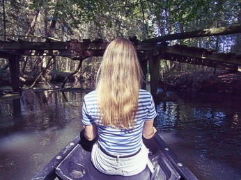 Eine junge Frau sitzt in einem Kanu, treibt auf einem ruhigen Fluss unter einer alten Holzbrücke. Das umgebende dichte Grün vermittelt Ruhe und Gelassenheit.