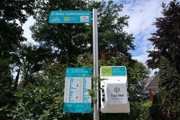 E-Bike-Ladestation in einem grünen, parkähnlichen Umfeld, umgeben von Bäumen und Sträuchern.