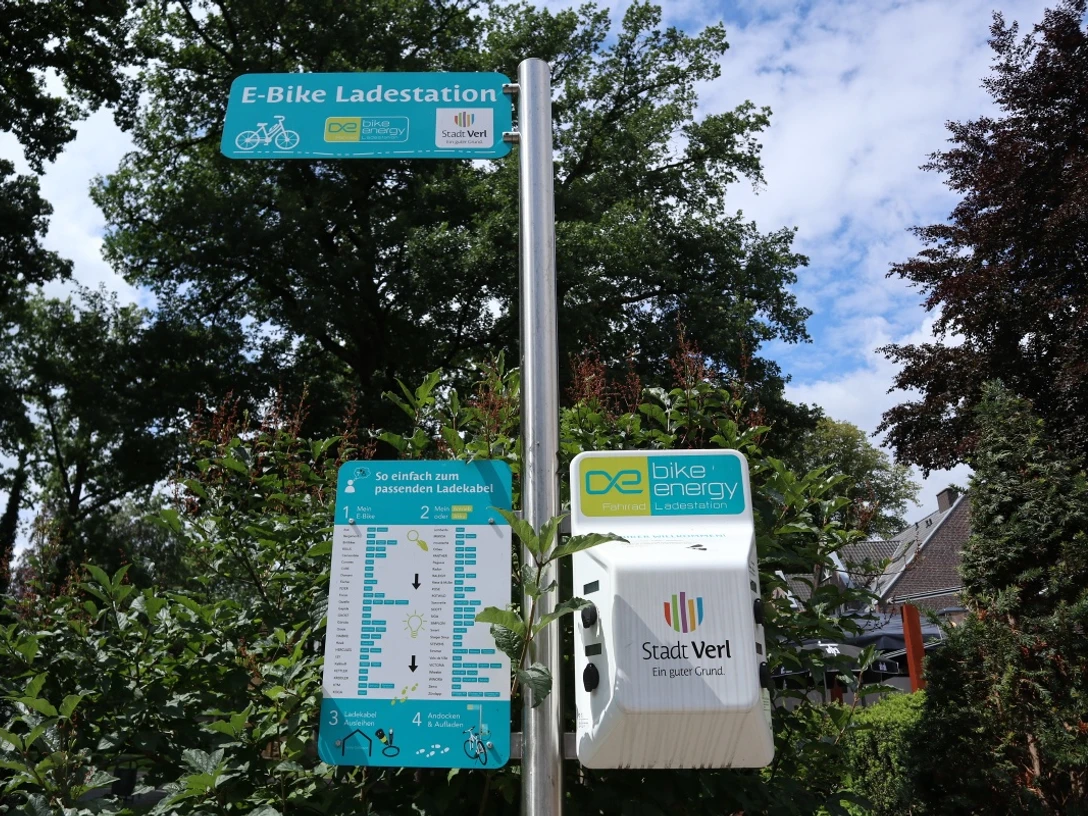 Infoschild an der E-Bike Ladestation E-Bike-Ladestation in einem grünen, parkähnlichen Umfeld, umgeben von Bäumen und Sträuchern.
