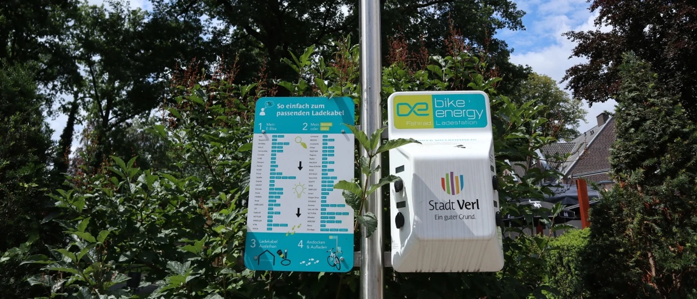 Infoschild an der E-Bike Ladestation E-Bike-Ladestation in einem grünen, parkähnlichen Umfeld, umgeben von Bäumen und Sträuchern.