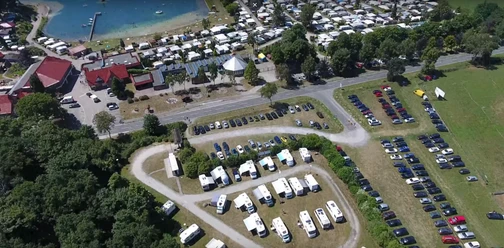 stellplatz_ferienpark Ein großer Parkplatz vor einem Ferienpark, mit geparkten Wohnmobilen und Blick auf einen See im Hintergrund.