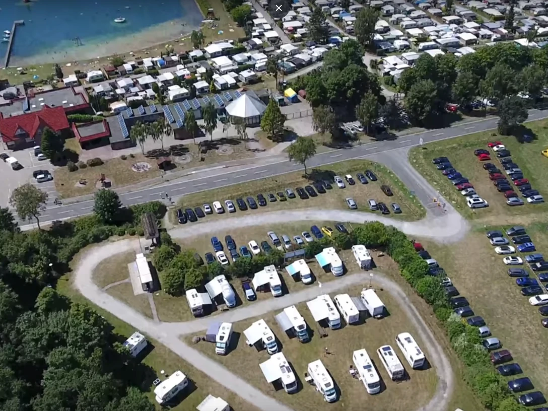 stellplatz_ferienpark Ein großer Parkplatz vor einem Ferienpark, mit geparkten Wohnmobilen und Blick auf einen See im Hintergrund.