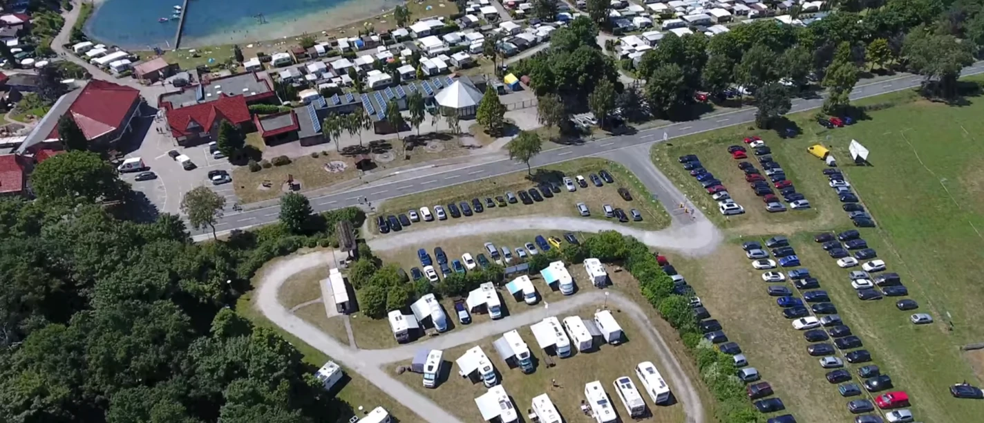 stellplatz_ferienpark Ein großer Parkplatz vor einem Ferienpark, mit geparkten Wohnmobilen und Blick auf einen See im Hintergrund.