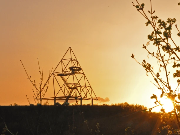 Tetraeder Bottrop <p>Ein eindrucksvolles Pyramidenkunstwerk bei Sonnenuntergang, mit blühenden Zweigen im Vordergrund.</p>