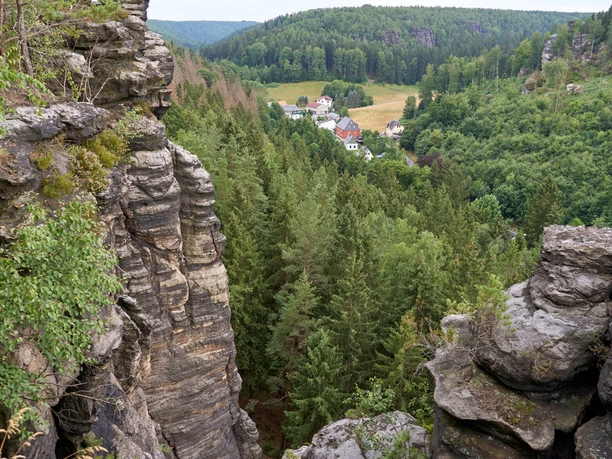 Ausblick vom Kanzelstein Felsformationen mit grünen Bäumen im Vordergrund, dahinter ein kleines Dorf mit roten und weißen Häusern, umgeben von dichtem Wald.