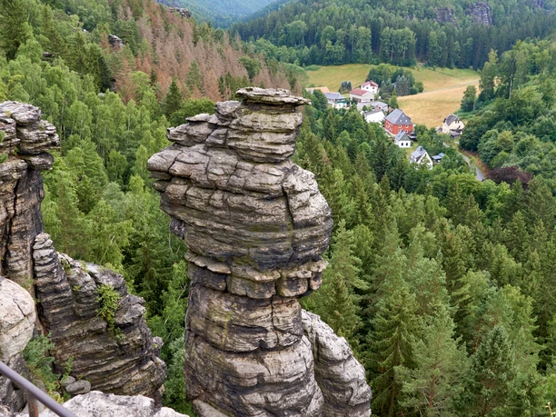 Aussichtspunkt Kanzelstein im Bielatal Felsformationen ragen über einen dichten, grünen Wald, im Hintergrund ein kleines Dorf mit weißen und roten Häusern.