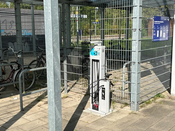 Fahrrad-Reparaturstation an einem sonnigen Tag, umgeben von Fahrradständern und Metallzäunen.