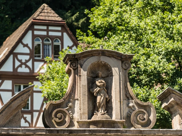 Altenberg Barocke Steinstatue mit filigranen Details vor einem historischen Fachwerkhaus und satter Naturkulisse.