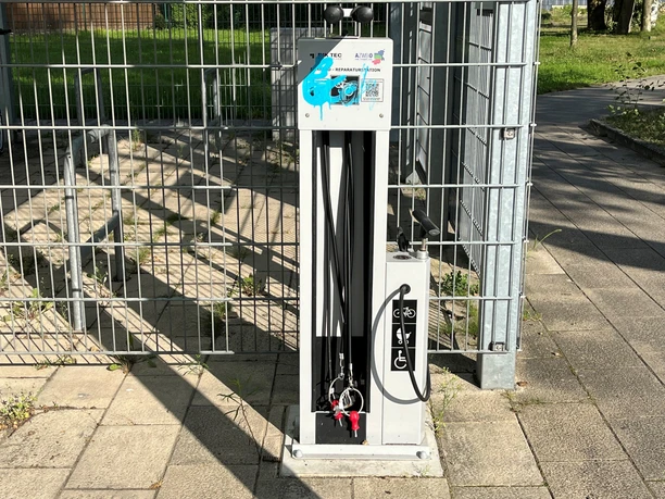 Fahrrad-Reparaturstation aus Metall mit Werkzeugen, Luftpumpe und QR-Code auf einem gepflasterten Weg.