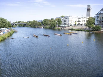 Drachenboot-Festival, Mülheim an der Ruhr Drachenboote wetteifern auf einem breiten Fluss, gesäumt von Bäumen und modernen Gebäuden.