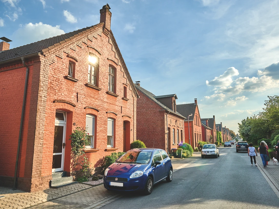 Die Mausegattsiedlung in Mülheim Rote Backsteinhäuser säumen eine ruhige Straße, während Menschen spazieren und Autos geparkt sind.