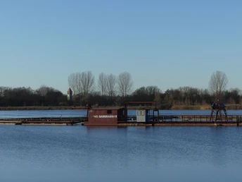 Blauer See mit Saugbagger im Vordergrund, Ufer mit Bäumen und einem Kirchturm im Hintergrund.