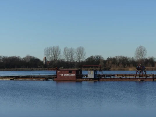 Baggersee zw. Anreppen und Delbrück Blauer See mit Saugbagger im Vordergrund, Ufer mit Bäumen und einem Kirchturm im Hintergrund.